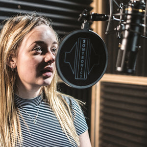 a girl with long hair on the left singing into a black Sontronics microphone on the right with a Sontronics popshield betwen her and the mic, in the background is studio acoustic treatment on the walls